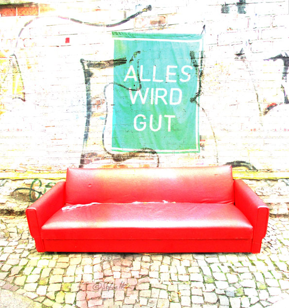 Rotes Ledersofa auf hellen Gehwegsteinen vor weißer Mauer mit günder Fahne, auf er in weißer Schrift steht: Alles wird gut.